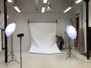 A wide shot of a professional photography studio where a woman is adjusting a large white paper backdrop. The setup includes multiple studio lights with white umbrellas and a black strobe light on stands.